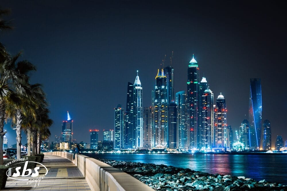 Una veduta notturna dello skyline della città con i grattacieli illuminati che si riflettono sull'acqua nelle meraviglie emiratine, una passerella di palme sulla sinistra e le rocce lungo il litorale in primo piano: un luogo stimolante per il turismo.