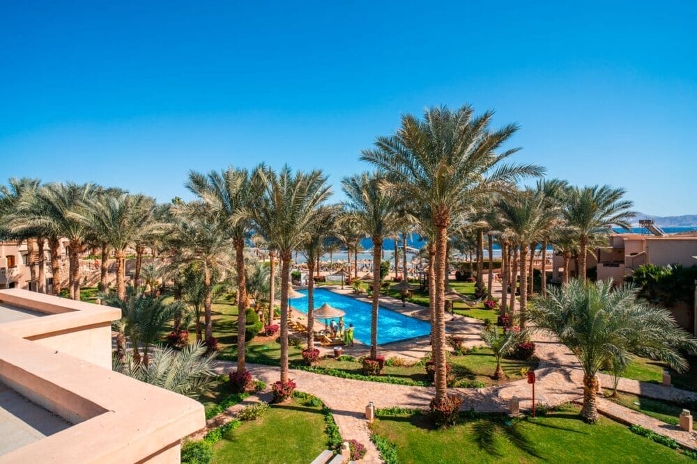 Un giardino del resort Bravo Tamra Beach con palme, una piscina sinuosa, prati verdi e sentieri, sullo sfondo di edifici beige e di un cielo azzurro. Il mare si staglia appena al di là dell'area del resort.