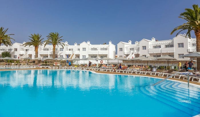 Una grande piscina all'aperto con acqua cristallina, circondata da lettini e ombrelloni, si trova di fronte agli edifici bianchi del resort Alpiclub Corralejo Village, incorniciata da palme sotto un cielo azzurro.