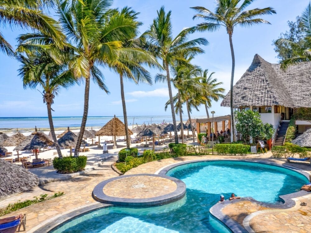 Un Beach Resort tropicale a Zanzibar con palme, piscina, capanne con tetto di paglia, sedie a sdraio e persone che si rilassano vicino all'acqua, affacciate su una spiaggia sabbiosa e sull'oceano blu chiaro sotto un cielo soleggiato.