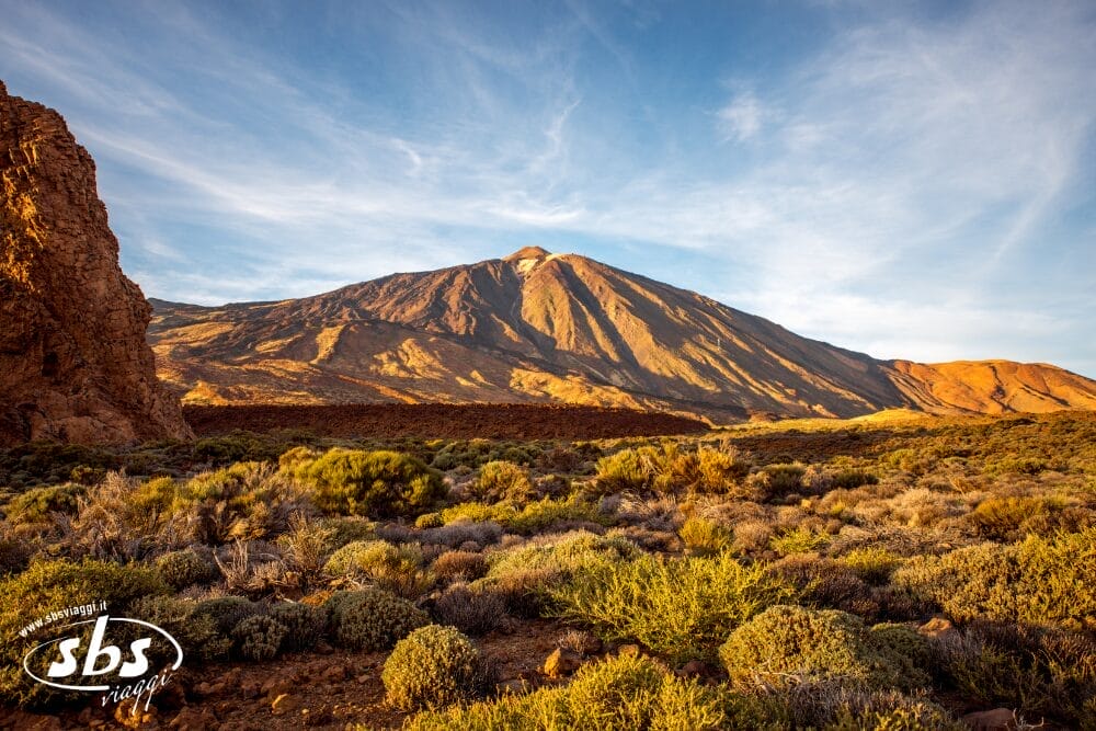Una veduta panoramica di un paesaggio arido e roccioso, con radi arbusti in primo piano e una montagna illuminata dal sole sotto un cielo azzurro con nuvole sottili, che cattura l'essenza di un tour "Meraviglie di Tenerife". La filigrana dell'immagine sbs è visibile nell'angolo in basso a sinistra.