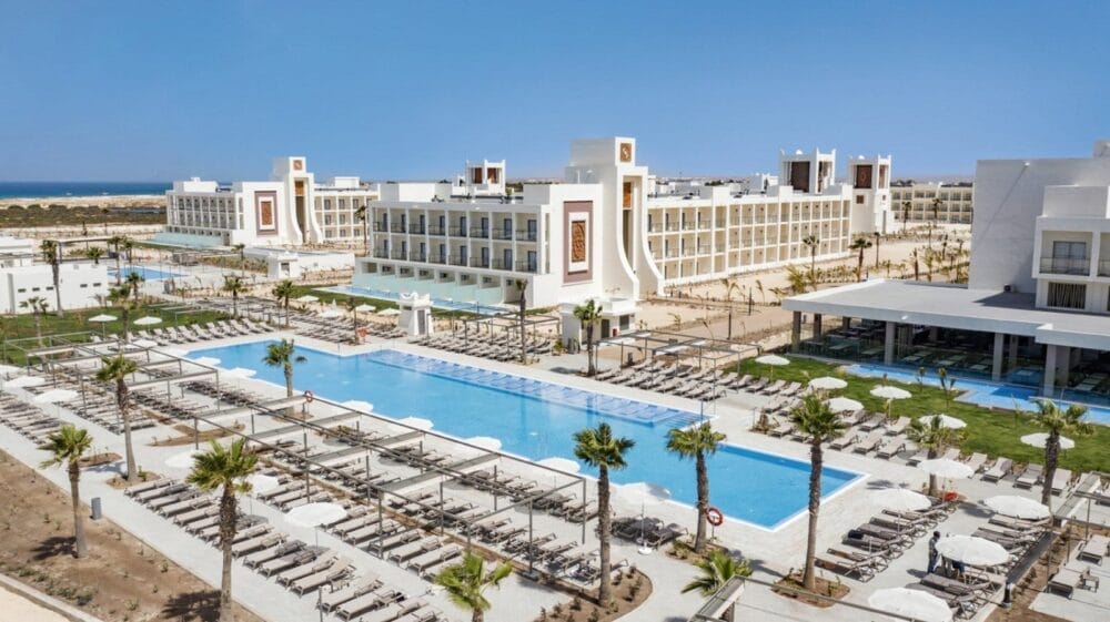 Il Riu Palace Santa Maria è un grande resort con diversi edifici bianchi circondati da palme, con una lunga piscina all'aperto fiancheggiata da sedie a sdraio e ombrelloni sotto un cielo azzurro e limpido. L'oceano è visibile in lontananza.