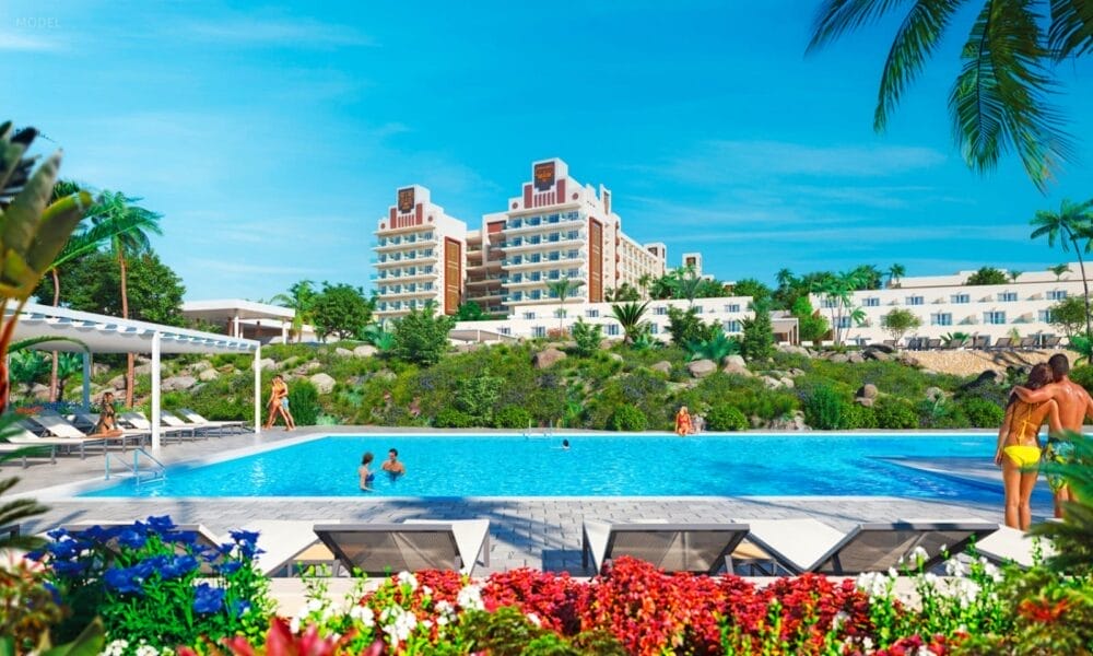 Il grande e moderno Riu Jambo Hotel si staglia sullo sfondo, circondato da palme e vegetazione lussureggiante. In primo piano, le persone si rilassano accanto a una piscina blu chiaro con sedie a sdraio e fiori colorati sotto un cielo blu brillante.