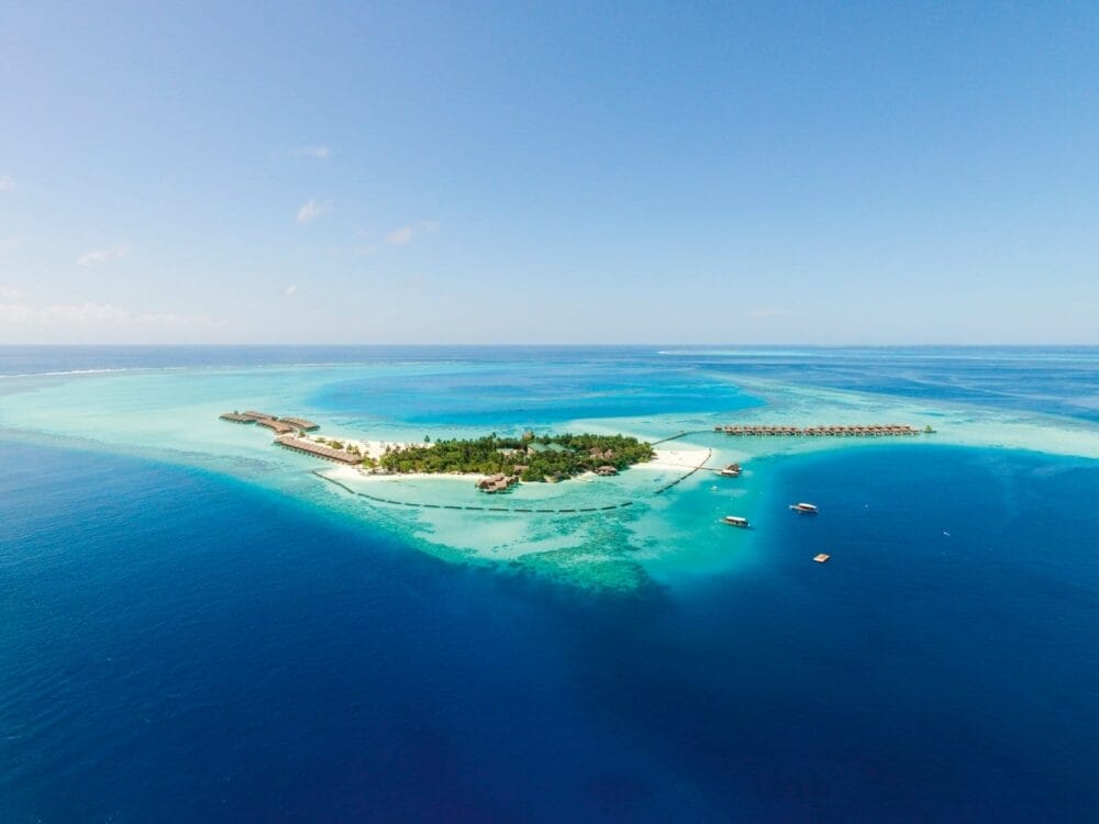 Vista aerea del Constance Moofushi, un resort di lusso alle Maldive circondato da acque turchesi, barriere coralline e oceano blu intenso. L'isola è caratterizzata da una vegetazione lussureggiante, aree di spiaggia, bungalow overwater e imbarcazioni vicine.