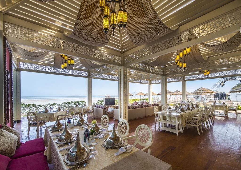 Un elegante ristorante all'aperto con decorazioni ornamentali, tavoli eleganti e cuscini si affaccia su una spiaggia con ombrelloni e l'oceano sullo sfondo. Le calde luci sospese creano un'atmosfera accogliente.