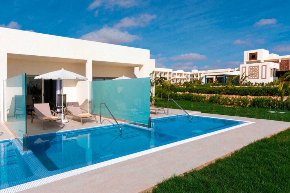 Moderna suite del resort Riu Palace Boavista con piscina privata, lettini, ombrellone e divisori in vetro. Un prato verde circonda la piscina, mentre altri edifici bianchi simili sono visibili sotto un cielo blu con nuvole sparse.