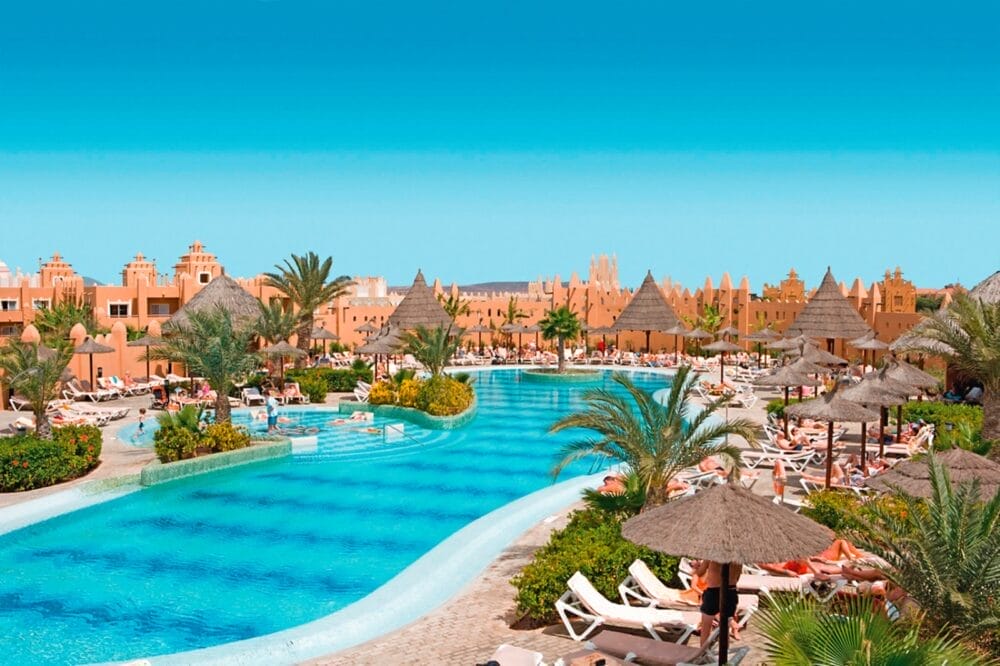 Grande piscina all'aperto del resort con acqua cristallina, circondata da palme, sedie a sdraio e ombrelloni di paglia. Le persone si rilassano a bordo piscina. Il cielo è limpido e sullo sfondo si vedono edifici dall'architettura unica.