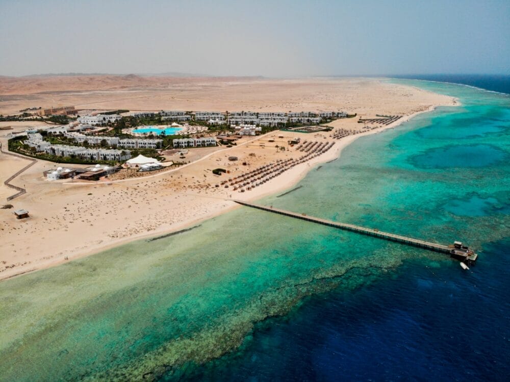 Veduta aerea del Gorgonia Beach Resort, uno splendido club marino con edifici bianchi, piscine e palme immerse nel deserto, accanto a un'acqua limpida e turchese e a un lungo molo che si protende nel mare.