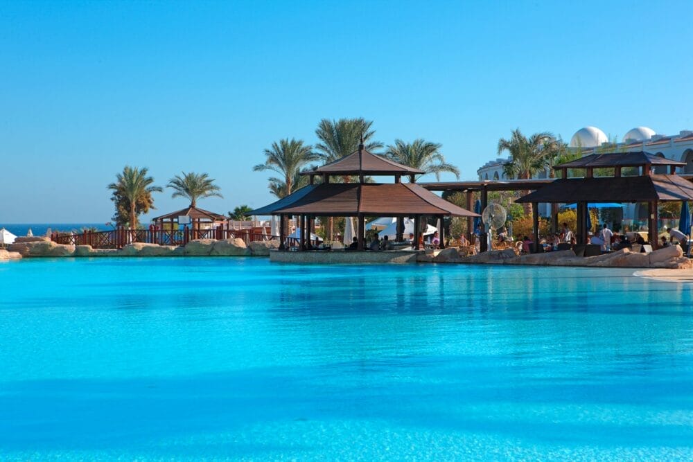 La grande piscina azzurra del Savoy Hotel, con padiglioni in legno a bordo piscina, palme e sedie a sdraio. Il cielo blu brillante incornicia gli edifici bianchi di Sharm El Sheikh e il mare in lontananza.