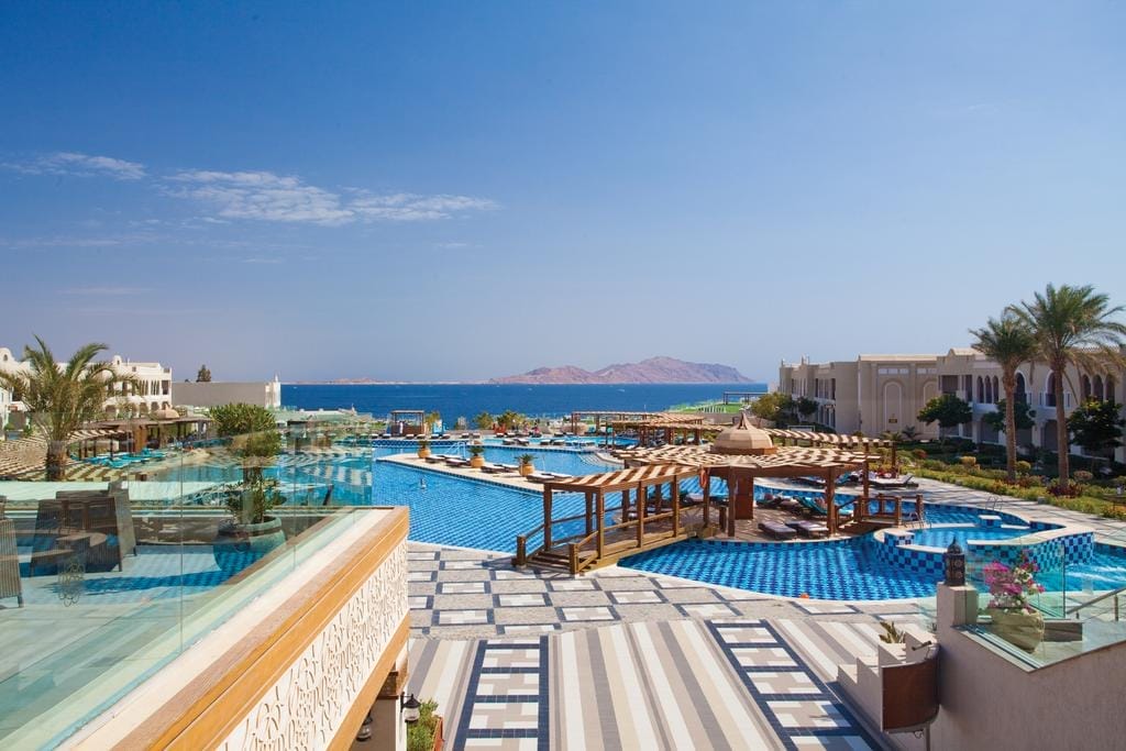 Il lussuoso resort Arabian Beach dispone di un'ampia piscina all'aperto con aree lounge ombreggiate, circondata da palme ed edifici eleganti, con vista sul mare blu e sulle montagne lontane sotto un cielo limpido.