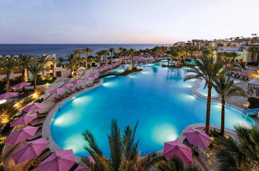 La grande e lussuosa piscina del Grand Rotana Resort è circondata da palme e ombrelloni rosa al tramonto, con sedie a sdraio ordinatamente disposte e l'oceano al di là. Questa scena tranquilla invita a rilassarsi presso il vicino Seaclub e la spa.