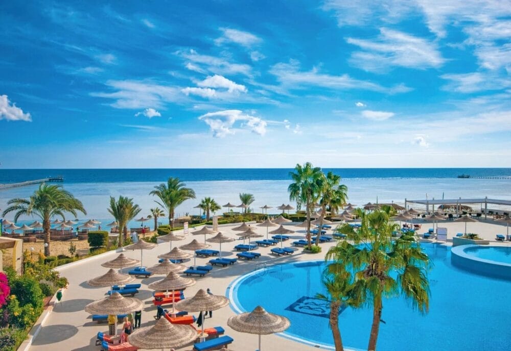 Un resort soleggiato con una grande piscina all'aperto, circondata da palme, lettini e ombrelloni di paglia. Al di là della piscina, la spiaggia di Blue Reef e il mare azzurro e calmo si estendono sotto un cielo luminoso con nuvole sparse.