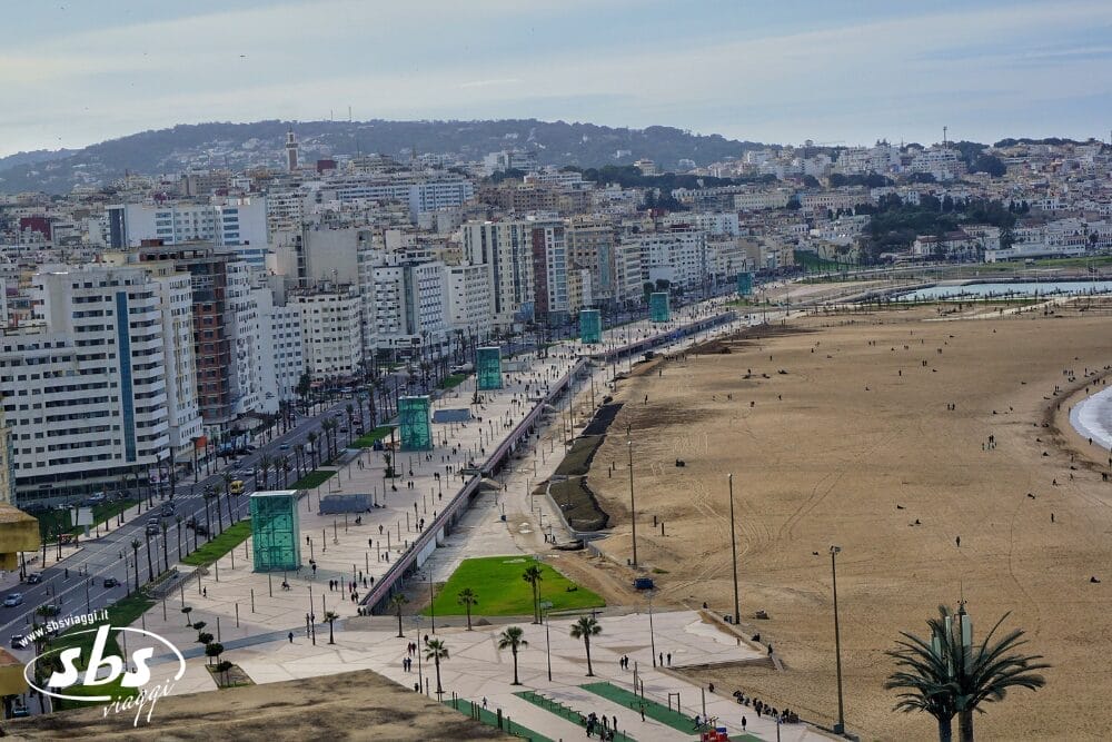 Veduta aerea del lungomare di una città del Marocco con alti edifici, un'ampia spiaggia sabbiosa, palme e persone che passeggiano sul lungomare. Sullo sfondo, sotto un cielo nuvoloso, si ergono colline ed edifici in stile Borghi Andalusi.