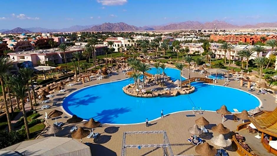 Una grande piscina del Parrotel Beach Resort con palme e lettini, circondata da edifici, ombrelloni di paglia e montagne lontane sotto un cielo azzurro e limpido a Sharm El Sheikh. Le persone si rilassano e nuotano intorno alla piscina.
