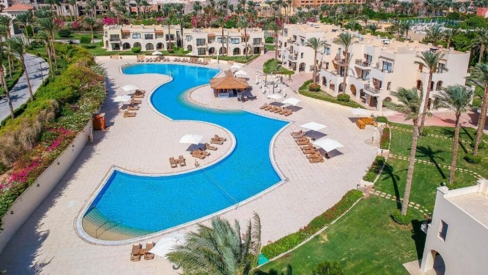 Vista aerea di una grande e sinuosa piscina all'aperto del Cleopatra Luxury Resort, circondata da lettini, ombrelloni, palme ed edifici del resort color crema in un giardino paesaggistico.