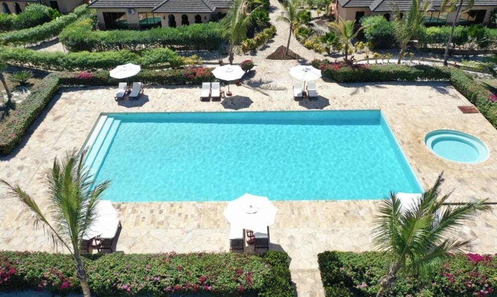 Vista aerea di una piscina esterna rettangolare con una piccola vasca idromassaggio rotonda nelle vicinanze, circondata da piastrelle in pietra, sedie a sdraio, ombrelloni bianchi, palme, verde paesaggistico e un tocco di eleganza automatica Bozza.