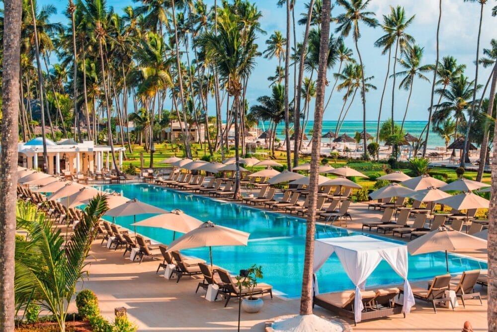 Una lussuosa piscina in vero stile Seaclub, circondata da sedie a sdraio e ombrelloni, con alte palme, eleganti cabanas e l'oceano che scintilla sullo sfondo sotto un cielo luminoso e soleggiato: una fuga da sogno.