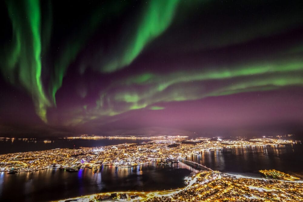 Veduta aerea di una città illuminata di notte a Capo Nord, con l'aurora boreale verde che vortica nel cielo stellato e si riflette leggermente sull'acqua che circonda la città.