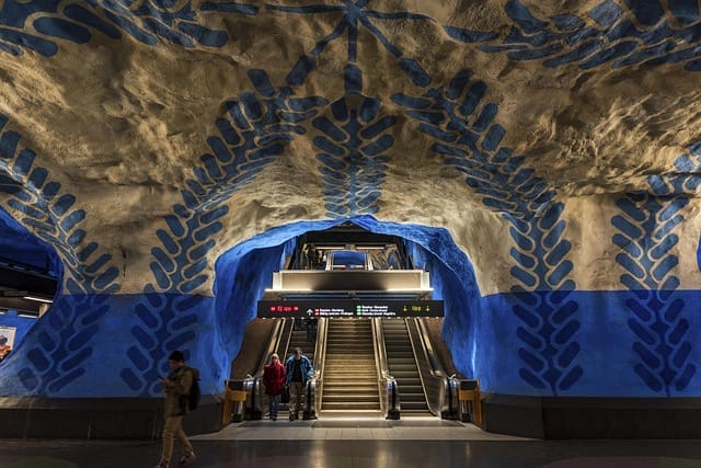 Le persone camminano vicino alle scale mobili in una stazione della metropolitana sotterranea con motivi artistici blu e beige dipinti sul soffitto e sulle pareti rocciose, creando un aspetto cavernoso che ricorda un capolavoro di Bozza automatico.