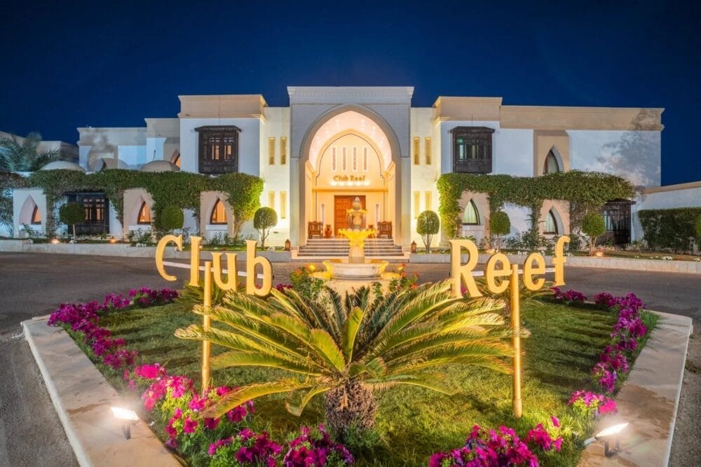 L'ingresso del Reef Beach Resort illuminato di notte, con una fontana centrale, cespugli curati, piante di palma e vivaci aiuole, con la scritta "Club Reef" sul prato di fronte a un elegante edificio bianco.