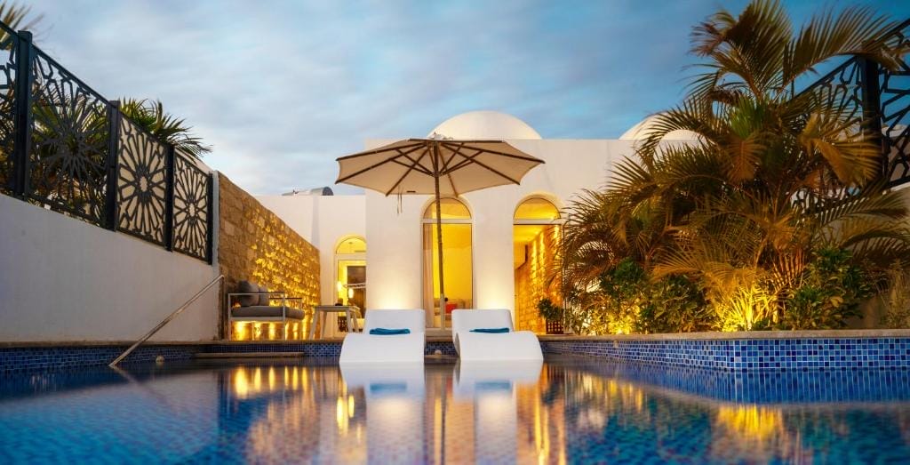 Una lussuosa scena a bordo piscina al tramonto del Fort Arabesque Resort mostra due sedie a sdraio bianche parzialmente immerse in una piscina dalle piastrelle blu, con un grande ombrellone tra di loro. Le luci calde illuminano le ville della spa, un edificio bianco e le palme lussureggianti sullo sfondo.