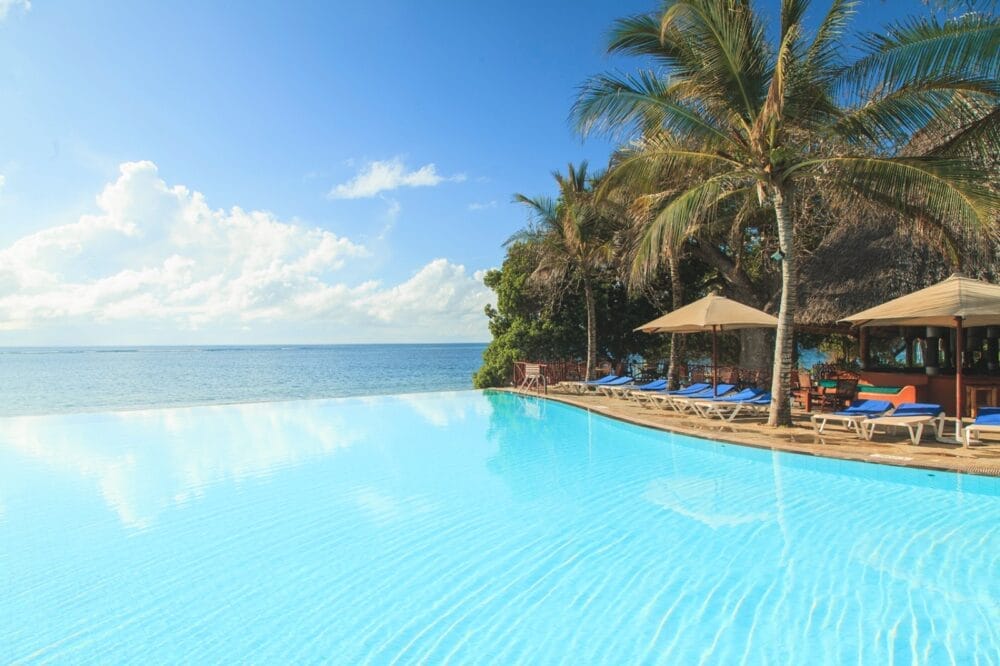 La grande piscina a sfioro del Seaclub si affaccia sull'oceano sotto un cielo azzurro e limpido, con palme, lettini e ombrelloni a bordo piscina che creano un'atmosfera tropicale e rilassante in stile Kole Kole.