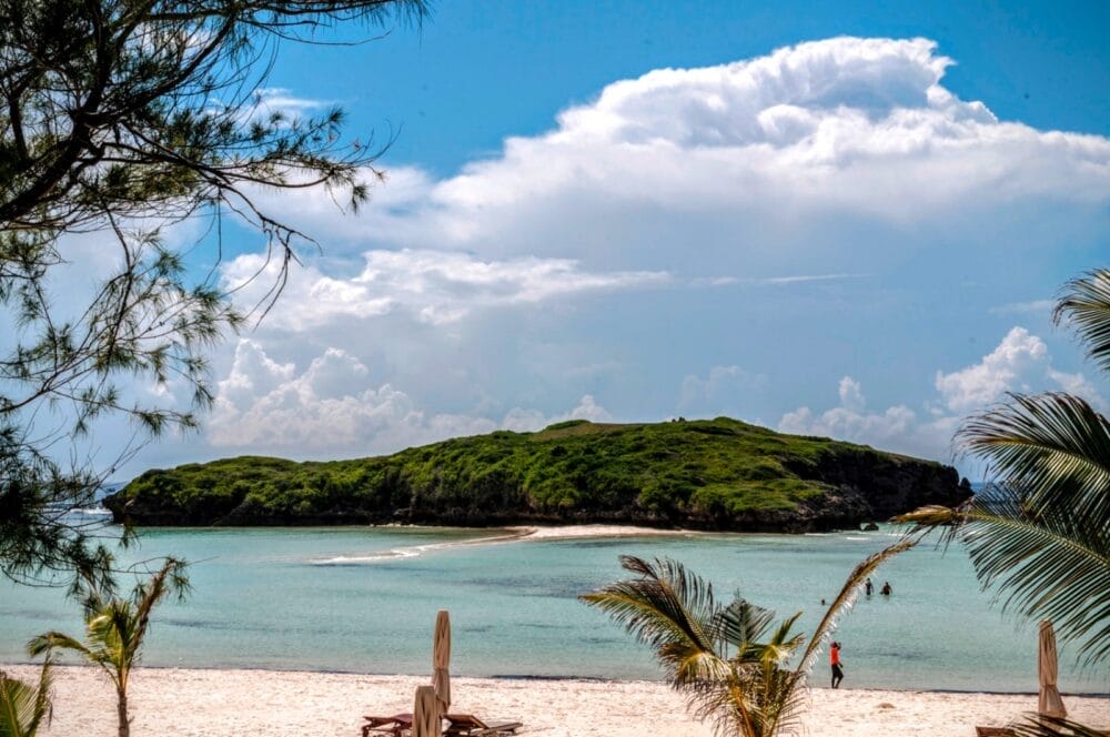 Una piccola isola verde e lussureggiante si trova al largo di una spiaggia sabbiosa con acqua turchese. Palme e sedie da spiaggia accolgono gli ospiti del Seaclub 7 Island Resort in primo piano, sotto un cielo parzialmente nuvoloso.
