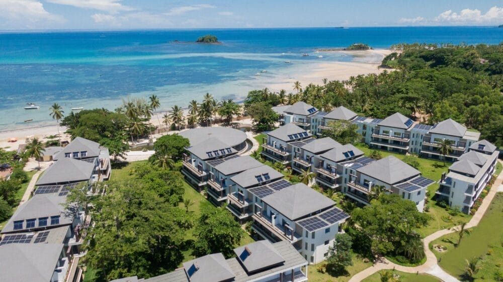 Vista aerea dell'Andilana Resort, con edifici bianchi, palme, una rilassante spa e pannelli solari sui tetti, che si affaccia su una spiaggia sabbiosa e un oceano turchese punteggiato da piccole isole in lontananza.