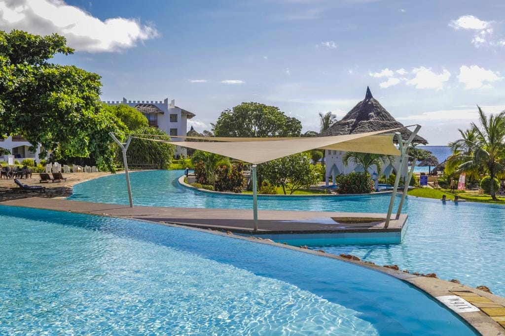 Ampia piscina all'aperto con bordi curvi presso il Royal Zanzibar Beach Resort, circondata da palme ed edifici bianchi. Una passerella ombreggiata attraversa la piscina sotto un cielo blu con nuvole sparse. Le persone si rilassano a bordo piscina.
