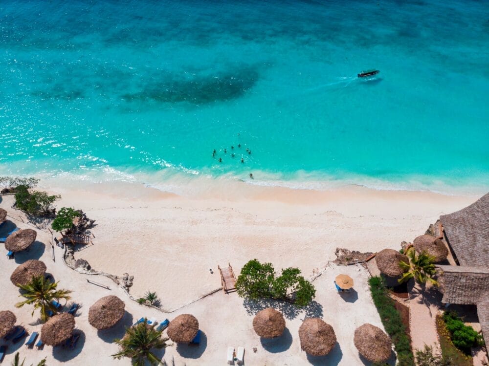 Vista aerea del Seaclub Sandies Baobab a Zanzibar: uno splendido resort sulla spiaggia con acqua turchese, ombrelloni di paglia e sedie a sdraio. Le persone nuotano vicino alla riva mentre una piccola barca galleggia sul mare calmo e trasparente che incontra la spiaggia sabbiosa.