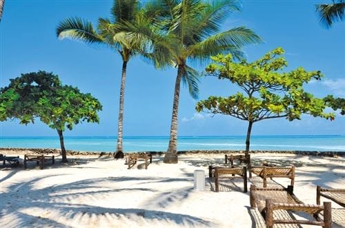 Scena soleggiata sulla spiaggia del Seaclub Diamond Mapenzi Beach con palme, sabbia bianca e sedie di vimini di fronte a un oceano blu e calmo. Il cielo limpido e luminoso rende l'atmosfera rilassante e tropicale.