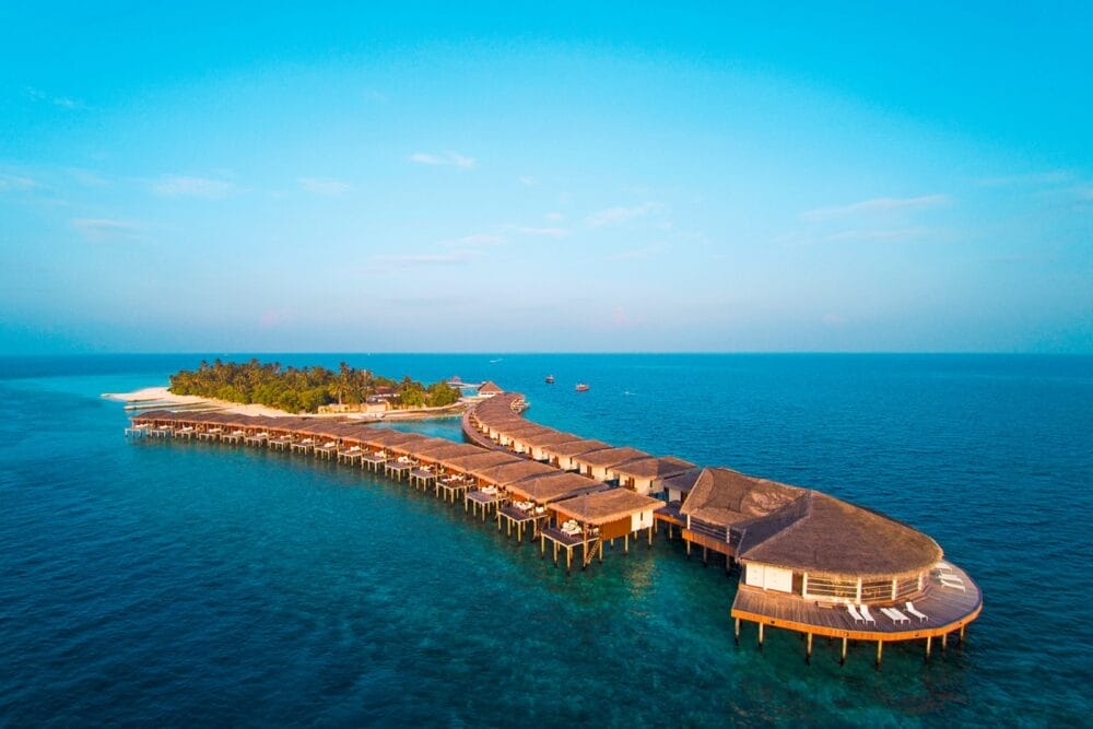Vista aerea del Seaclub Dhiggiri, un resort alle Maldive con ville overwater che si estendono su un'isola tropicale, circondata da un oceano cristallino, da una vegetazione lussureggiante e da spiagge sabbiose sotto un cielo limpido e luminoso.