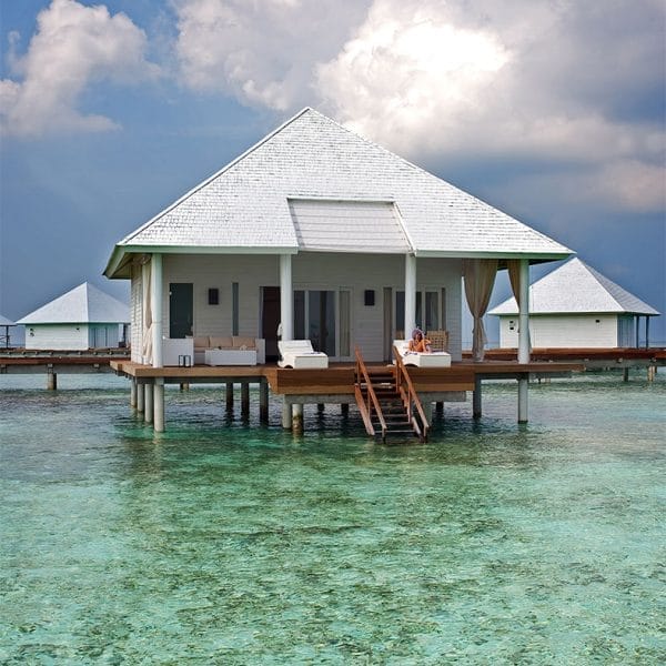 Una Water Villa bianca in stile Seaclub con terrazza si trova sopra l'acqua limpida e turchese di Athuruga Beach, sotto un cielo parzialmente nuvoloso, con un altro bungalow simile visibile sullo sfondo.