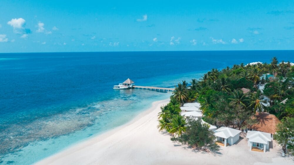 Vista aerea del Sandies Bathala Resort: una spiaggia tropicale con sabbia bianca, acqua turchese, un molo di legno con una capanna dal tetto di paglia e diverse ville bianche circondate da palme lussureggianti. Scoprite il paradiso in questa destinazione Seaclub.