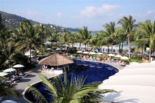Un resort tropicale a Kamala Beach con una grande piscina curva, circondata da palme, lettini con cuscini colorati e ombrelloni bianchi. L'oceano e le verdi colline sono visibili sullo sfondo sotto un cielo azzurro.