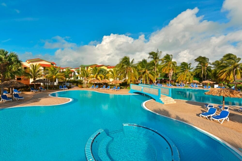 Grande piscina all'aperto con un piccolo ponte, circondata da sedie a sdraio blu, palme ed edifici tropicali sotto un cielo blu brillante all'Iberostar Tainos Seaclub.