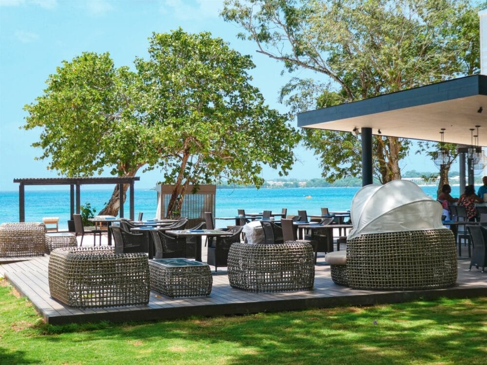 Patio del ristorante all'aperto con moderni mobili in vimini e ombra al Royalton Negril, affacciato su un oceano blu brillante, con grandi alberi che forniscono verde e un cielo limpido sopra.