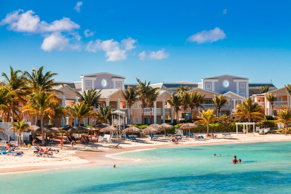 Resort Grand Palladium Jamaica fronte spiaggia con edifici dai colori pastello, palme e lettini sotto ombrelloni di paglia sulla sabbia bianca. Gli ospiti si rilassano e nuotano nell'oceano turchese sotto un cielo blu brillante con poche nuvole.