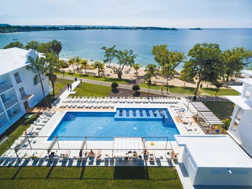Vista aerea del Seaclub Riu Negril, un resort all-inclusive della Giamaica con una grande piscina all'aperto, lettini, palme e ospiti che si rilassano. Il resort di Negril si trova accanto a una spiaggia sabbiosa e a un tranquillo oceano blu sotto un cielo limpido.