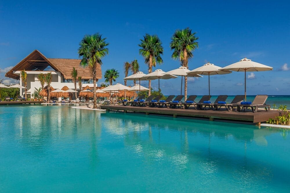 Una piscina del Searesort con sedie a sdraio e ombrelloni accanto all'acqua cristallina, circondata da palme. Un edificio con tetto di paglia e l'oceano sono visibili sullo sfondo sotto un cielo blu brillante all'Ocean Riviera Paradise.