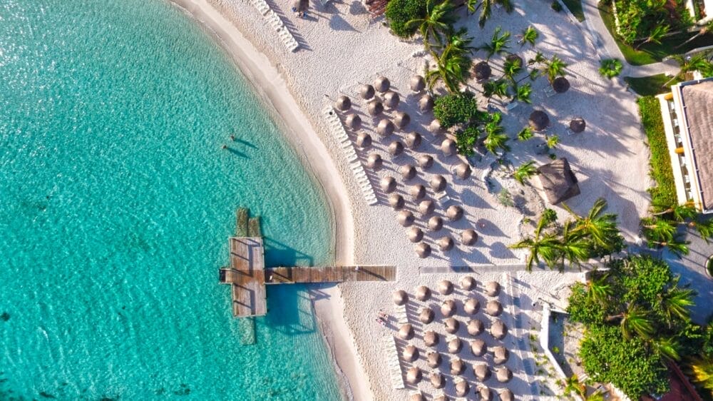 Veduta aerea di una spiaggia tropicale nei pressi di Luxury Akumal, con acque turchesi limpide, un molo di legno, sabbia bianca, file di ombrelloni di paglia e palme verdi.