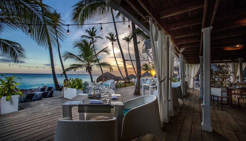 Il ristorante sulla spiaggia del Seaclub Catalonia Bayahibe offre tavoli e sedie bianche con vista sull'oceano al tramonto. Palme, luci e tende trasparenti creano un'atmosfera rilassata e tropicale al Gran Dominicus.