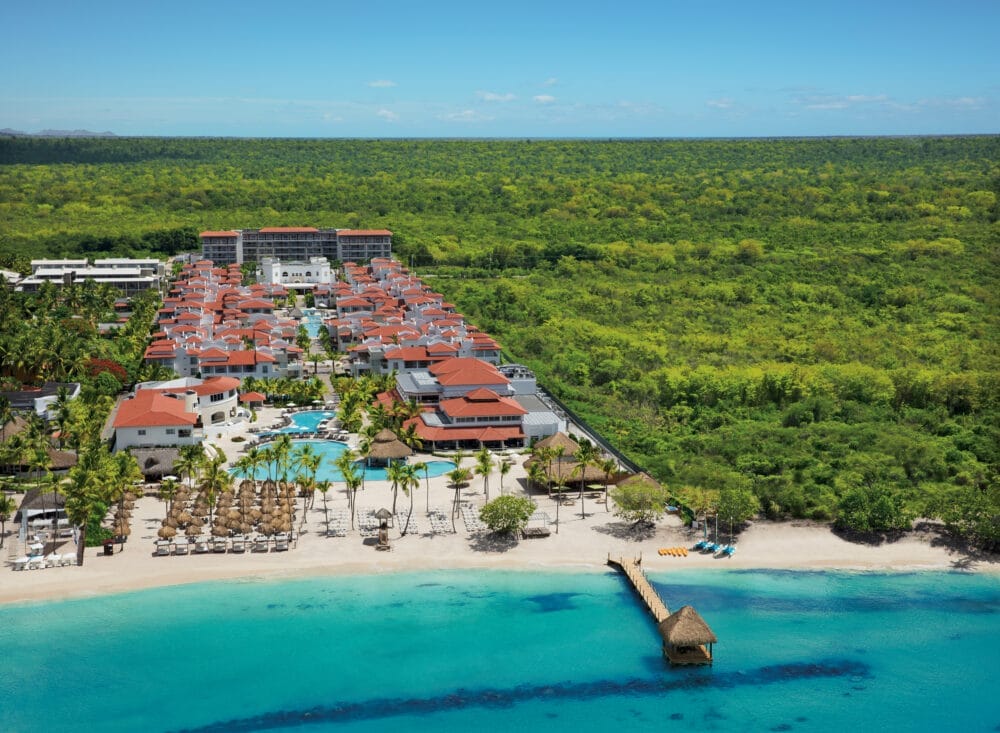 Vista aerea del Seaclub Style Dreams Dominicus a La Romana: resort con tetto rosso sulla spiaggia, piscina, palme, lettini da spiaggia, pontile in legno con capanna di paglia e sfondo di foresta lussureggiante. L'oceano azzurro e limpido scintilla in primo piano.