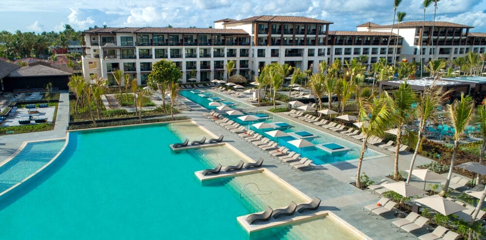 Vista aerea del lussuoso resort Lopesan Costa Bavaro con diverse piscine, lettini e palme, con un grande edificio a più piani sullo sfondo sotto un cielo parzialmente nuvoloso.