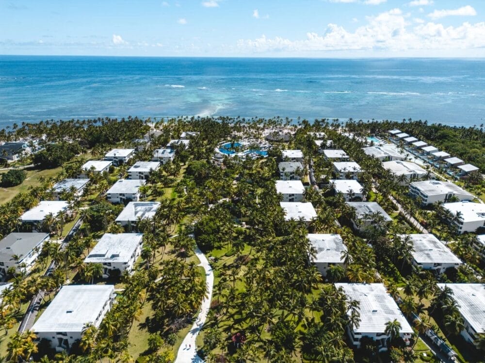Vista aerea del resort Catalonia Bavaro, con edifici dai tetti bianchi incastonati tra palme, sentieri tortuosi e un'area piscina centrale, il tutto affacciato sull'oceano blu di Bavaro Beach sotto un cielo parzialmente nuvoloso.