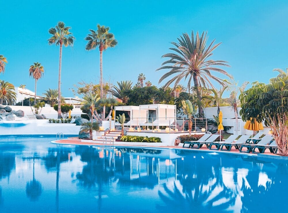 La tranquilla piscina all'aperto del Seaclub H10 Gran Tinerfe è circondata da sedie a sdraio, ombrelloni gialli, palme e vegetazione lussureggiante sotto un cielo blu brillante, con moderni edifici bianchi sullo sfondo.