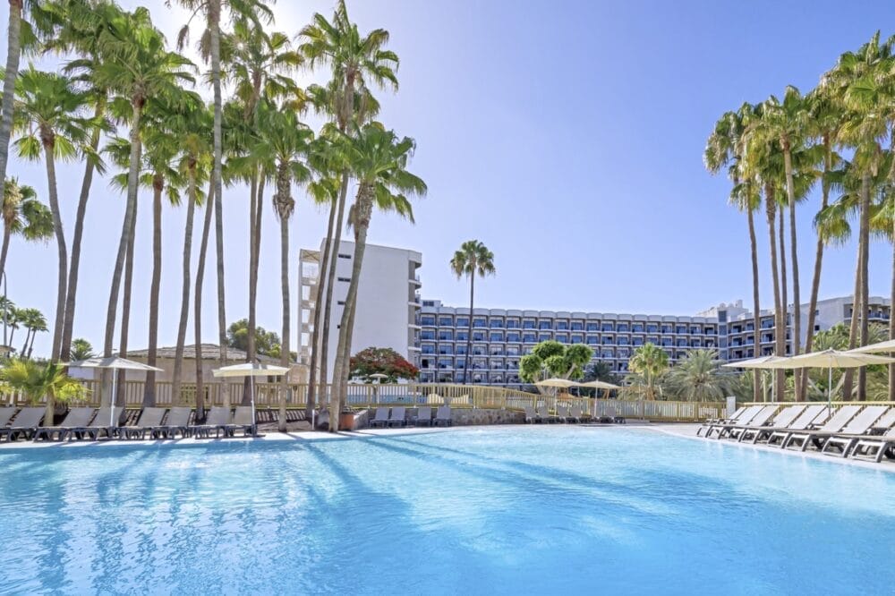 Una grande piscina all'aperto circondata da sedie a sdraio e alte palme, con il moderno hotel Seaclub Barcelò Margaritas che si staglia sullo sfondo sotto un cielo azzurro e limpido.