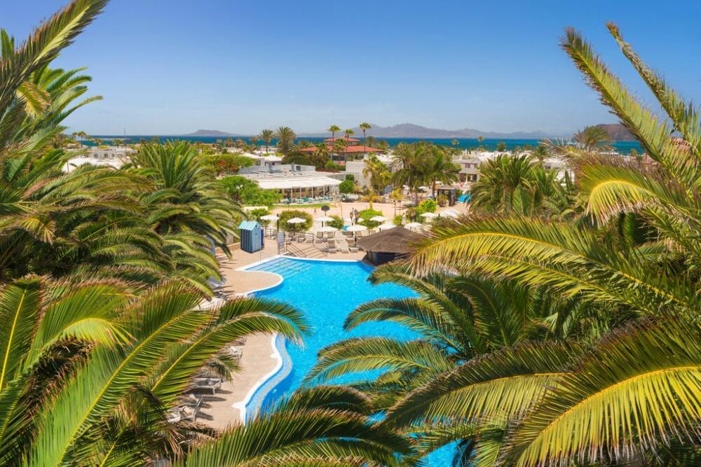 Una vista luminosa e soleggiata dell'Alua Suites Fuerteventura Searesort con una piscina blu sinuosa circondata da palme lussureggianti, lettini ed edifici bianchi. L'oceano e le montagne lontane sono visibili sotto un cielo azzurro e limpido.
