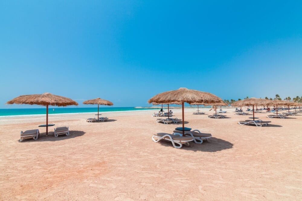 Ampia spiaggia sabbiosa del Seaclub Fanar con ombrelloni di paglia e sedie a sdraio bianche sotto un cielo azzurro e limpido. Il mare turchese è calmo sullo sfondo, creando una scena tranquilla e non affollata, con poche persone che si rilassano in lontananza.