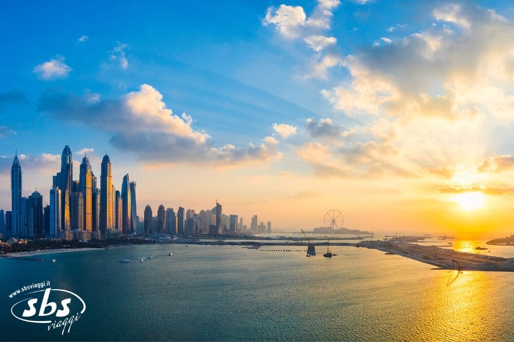 Una vista panoramica dello skyline di Dubai Marina con alti grattacieli, una baia tranquilla e il sole che tramonta, proiettando raggi dorati attraverso le nuvole sulla città e sull'acqua. Il logo di SBS Viaggi e Bozza automatica sono nell'angolo in basso a sinistra.